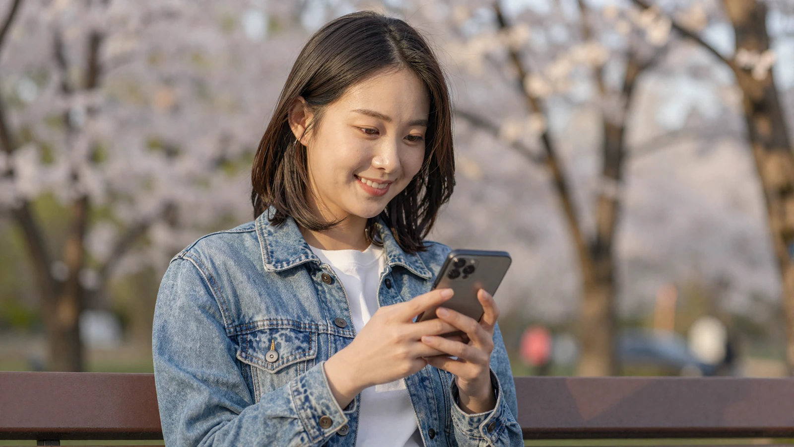 Young Asian woman smiling at her phone on a park bench - learn how to start a conversation on dating apps